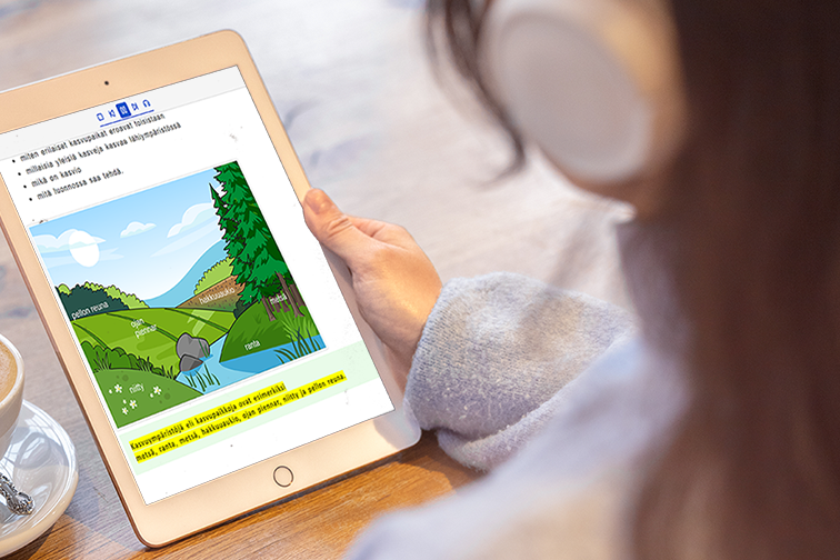 Henkilö, jolla on päässään valkoiset kuulokkeet, pitää käsissään tablet-laitetta, jossa on auki Celian ympäristöopin oppikirja. Näytöllä on kuva luonnosta, jossa on kasvien erilaisia kasvuympäristöjä, kuten metsä, ranta ja niitty. Kuvassa on tekstiä, joka selittää kasvien ja ympäristöjen eroja. Taustalla on puupintainen pöytä, jolla on kahvikuppi. 